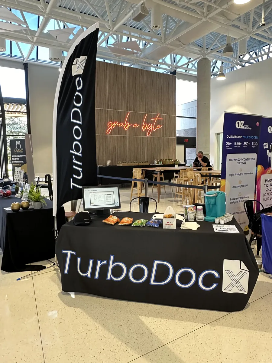 TurboDocx conference booth with a Goldcoast Marketing Group branded table cover, feather flag, and event swag
